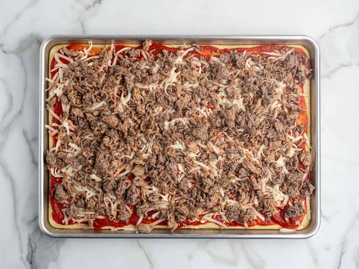 Rectangular pizza dough on a baking sheet topped with tomato sauce, shredded cheese, and cooked ground beef, ready to be bakedโjust like a classic Rectangle School Pizza. Marble countertop underneath.