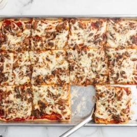 A sheet pan of rectangular, cheesy sausage pizza slicesโjust like classic Rectangle School Pizzaโwith one slice removed; a spatula rests in the empty space on a marble countertop.