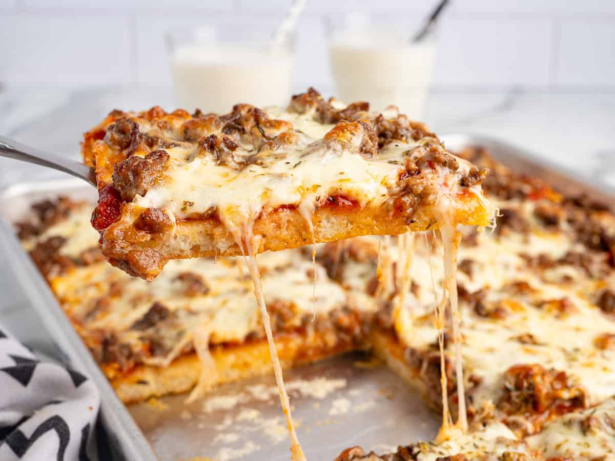 A spatula lifts a cheesy, beef-topped slice of Rectangle School Pizza from a pan, with gooey cheese stretching. Two glasses of milk are in the background.