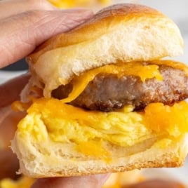A close-up of a hand holding one of the Sausage Egg and Cheese Breakfast Sliders, featuring scrambled eggs, cheddar cheese, and a savory sausage patty inside a soft, golden bun.