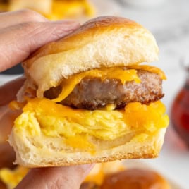 A hand holds one of the Sausage Egg and Cheese Breakfast Sliders, featuring a sausage patty, scrambled eggs, and melted cheddar cheese on a soft bun.