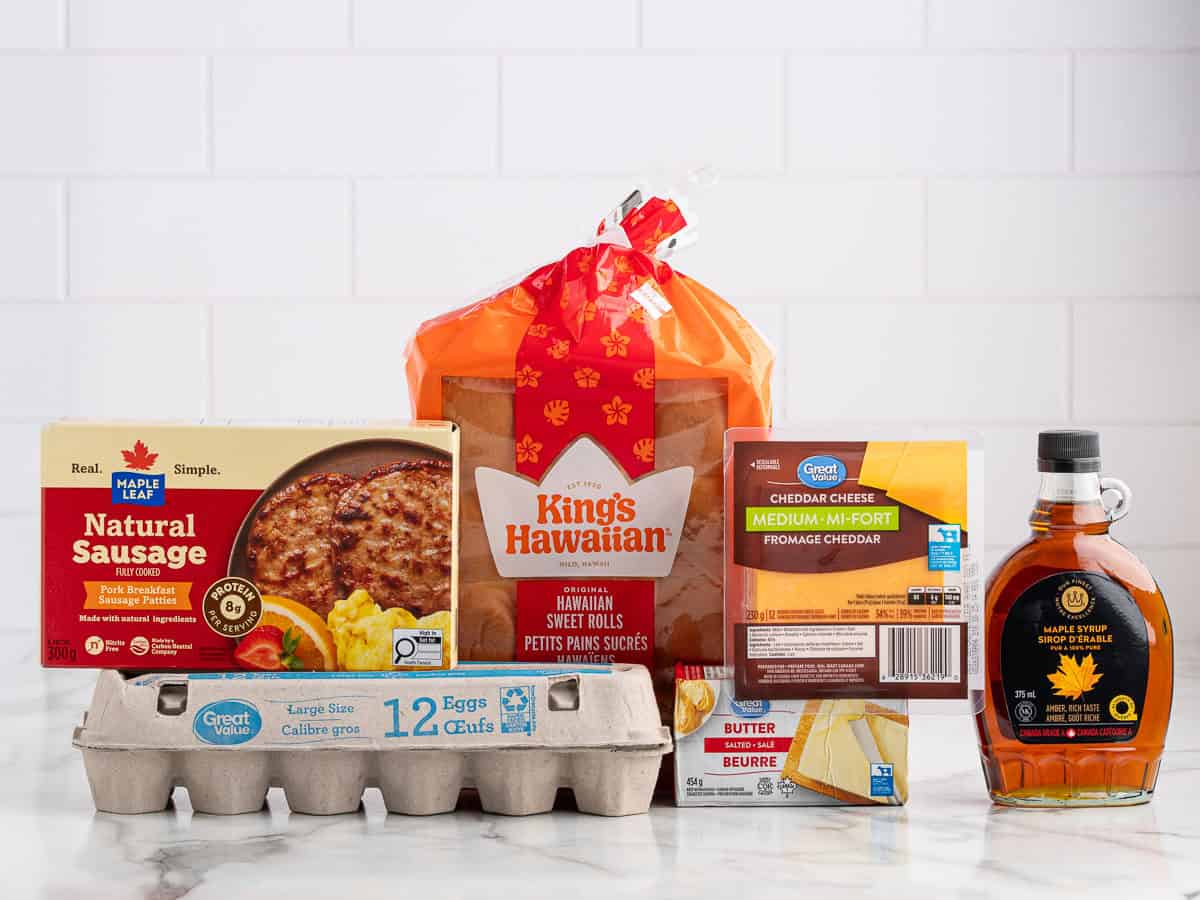 A variety of breakfast items on a white counter, including eggs, sausage, Kings Hawaiian sweet rolls, cheddar cheese, butter, and maple syrup—perfect for assembling delicious Sausage Egg and Cheese Breakfast Sliders.