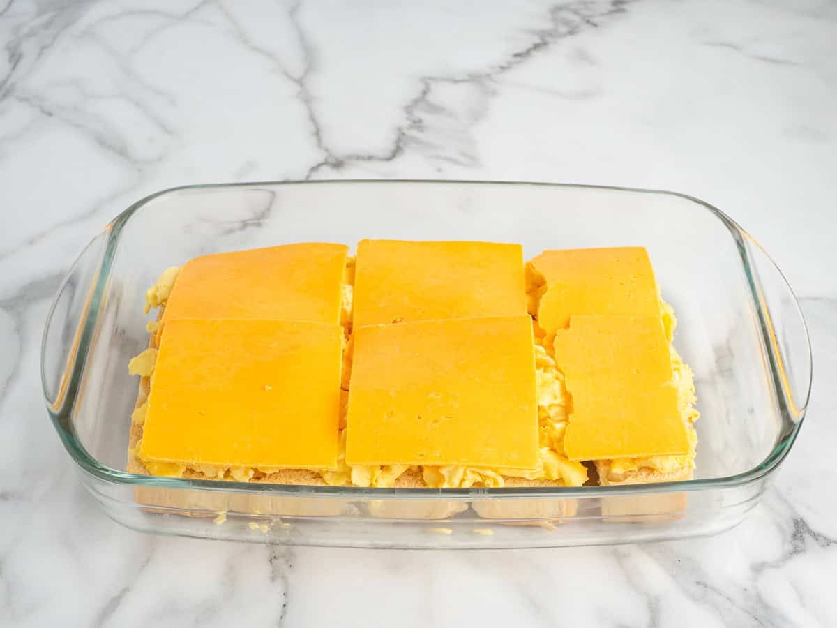A glass baking dish with six slices of cheddar cheese on top of a layered casserole, reminiscent of delicious Sausage Egg and Cheese Breakfast Sliders, set on a white marble surface.