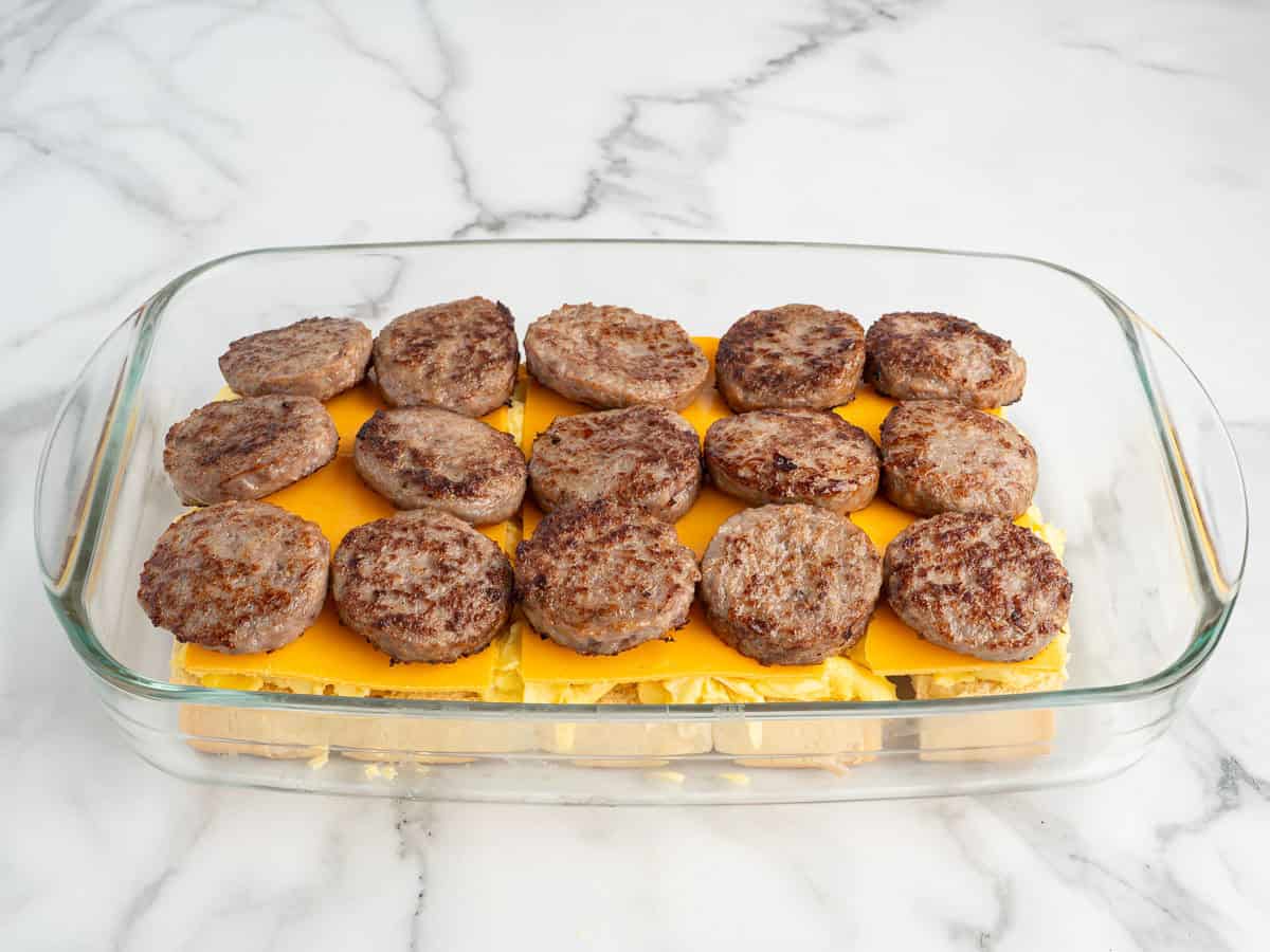 A glass baking dish containing layers of cooked sausage patties, cheddar cheese slices, and scrambled eggs on a marble countertop—perfect for preparing delicious Sausage Egg and Cheese Breakfast Sliders.