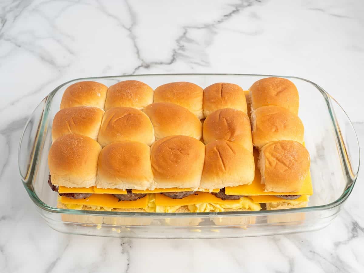A glass baking dish filled with assembled Sausage Egg and Cheese Breakfast Sliders, made with square bread rolls, cheese slices, and savory patties, sits on a white marble surface.