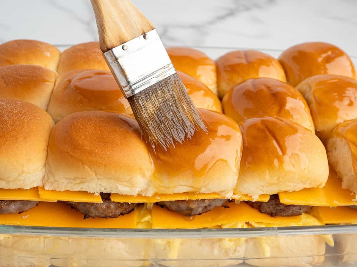 A brush is spreading melted butter on top of a row of Sausage Egg and Cheese Breakfast Sliders with golden buns in a glass baking dish.