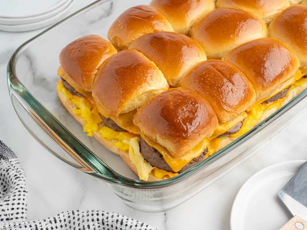 A glass baking dish filled with golden brown Sausage Egg and Cheese Breakfast Sliders made with buns, scrambled eggs, melted cheese, and savory sausage patties rests on a marble countertop.