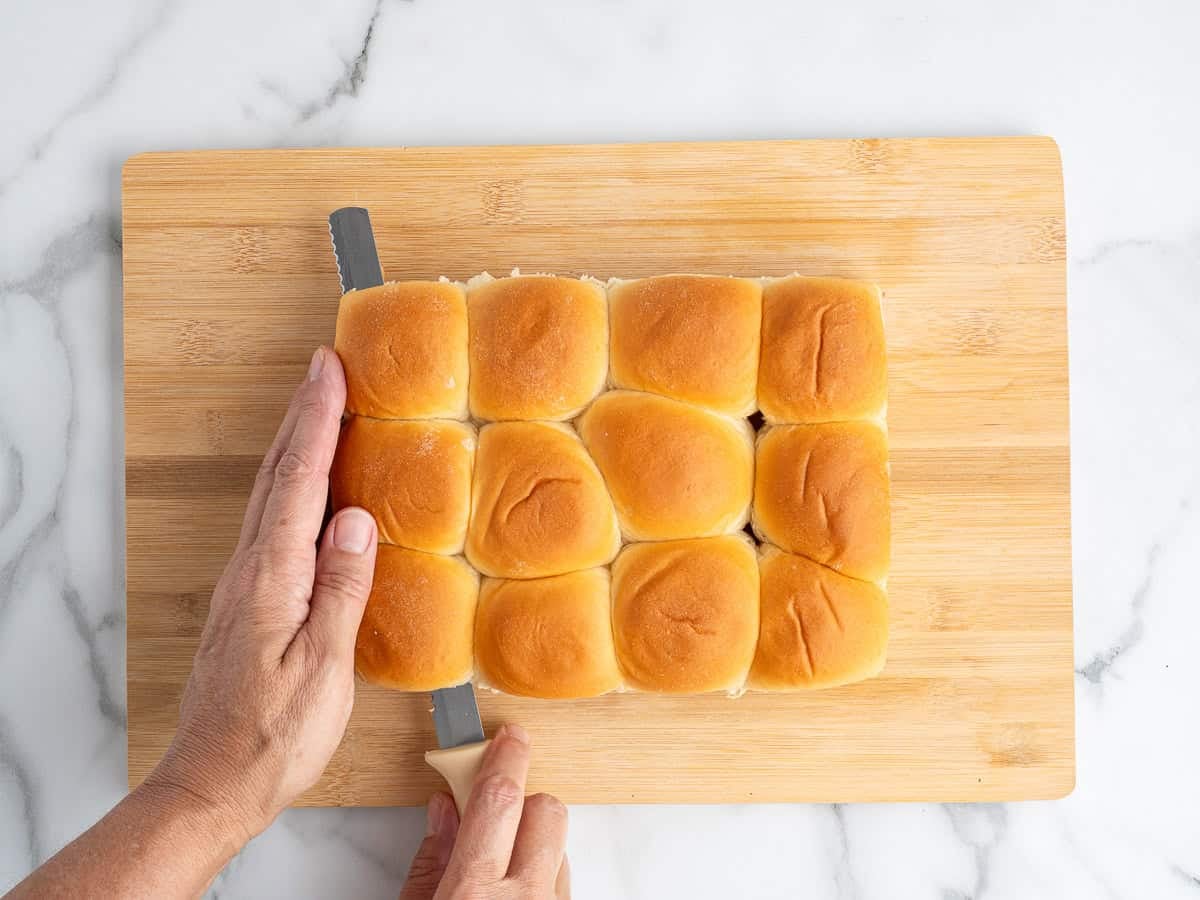 Two hands slice a group of twelve dinner rolls in half horizontally on a wooden cutting board, preparing the base for delicious Sausage Egg and Cheese Breakfast Sliders.