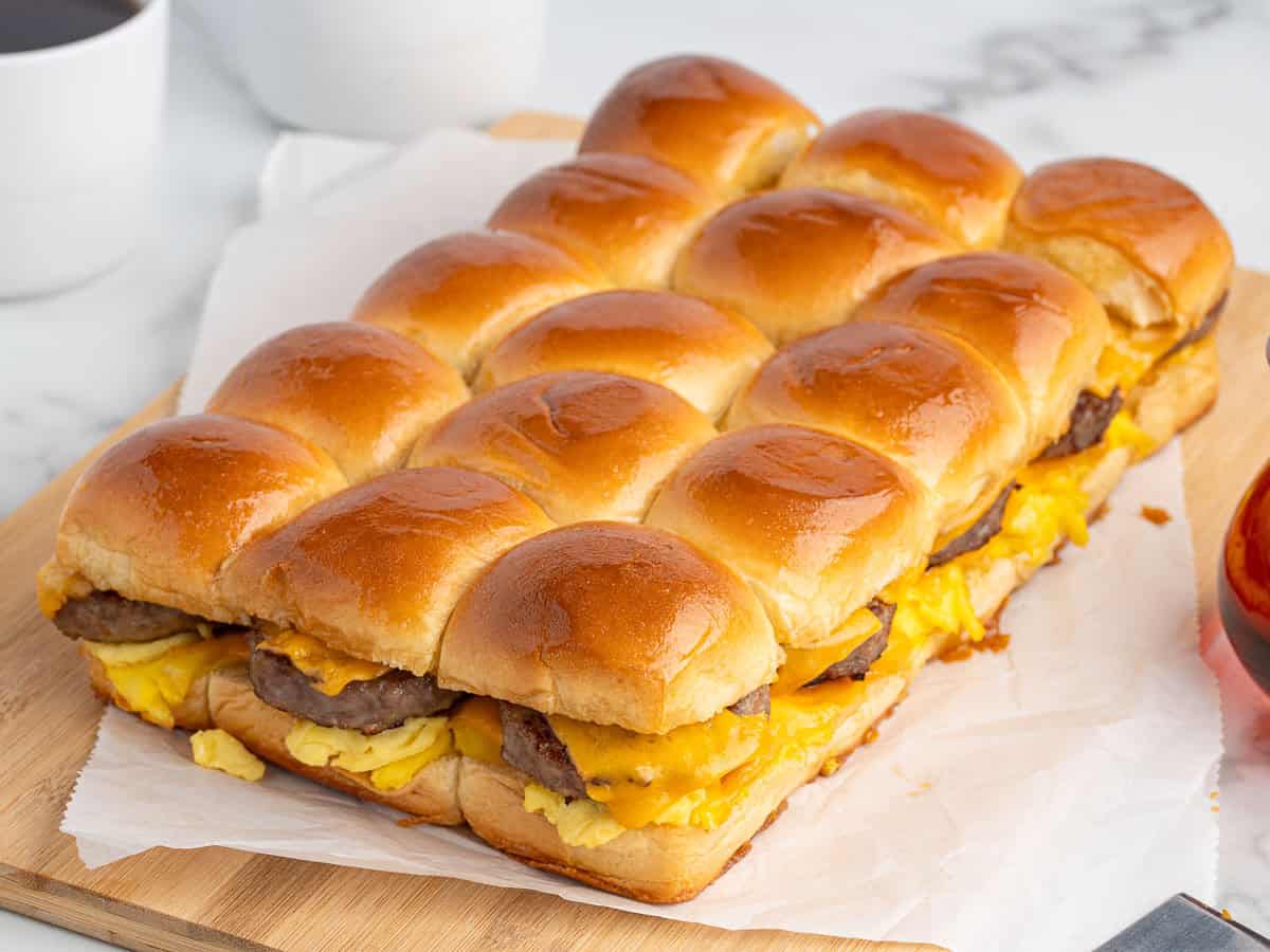 A tray of Sausage Egg and Cheese Breakfast Sliders made with golden buns, eggs, sausage, and melted cheese is arranged neatly on parchment paper atop a wooden board.