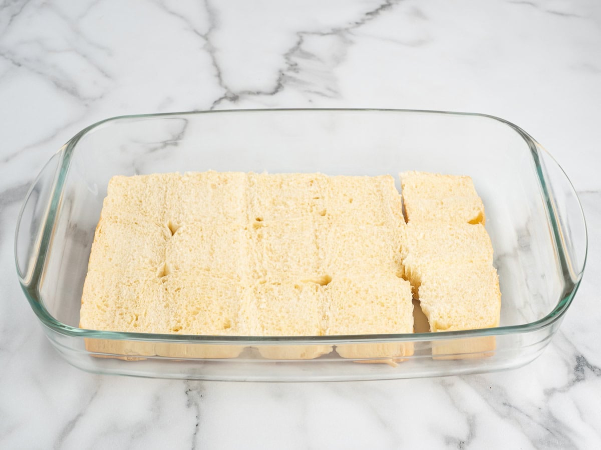 A glass baking dish with slices of white bread arranged in a single layer, covering the bottom, on a white marble surface—perfect as the base for assembling Sausage Egg and Cheese Breakfast Sliders.