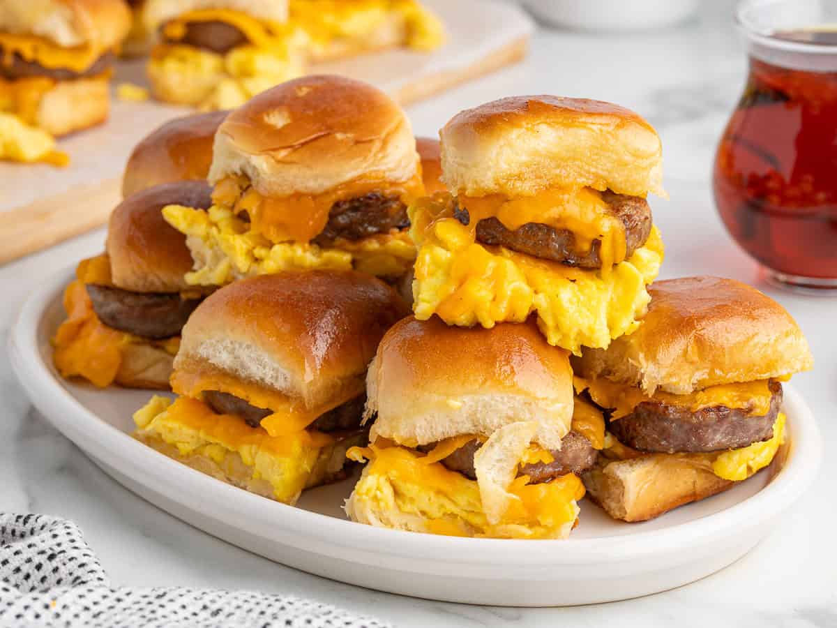 A plate of Sausage Egg and Cheese Breakfast Sliders made with fluffy scrambled eggs, savory sausage patties, and melted cheddar cheese on soft buns, served with syrup in a small glass jar nearby.
