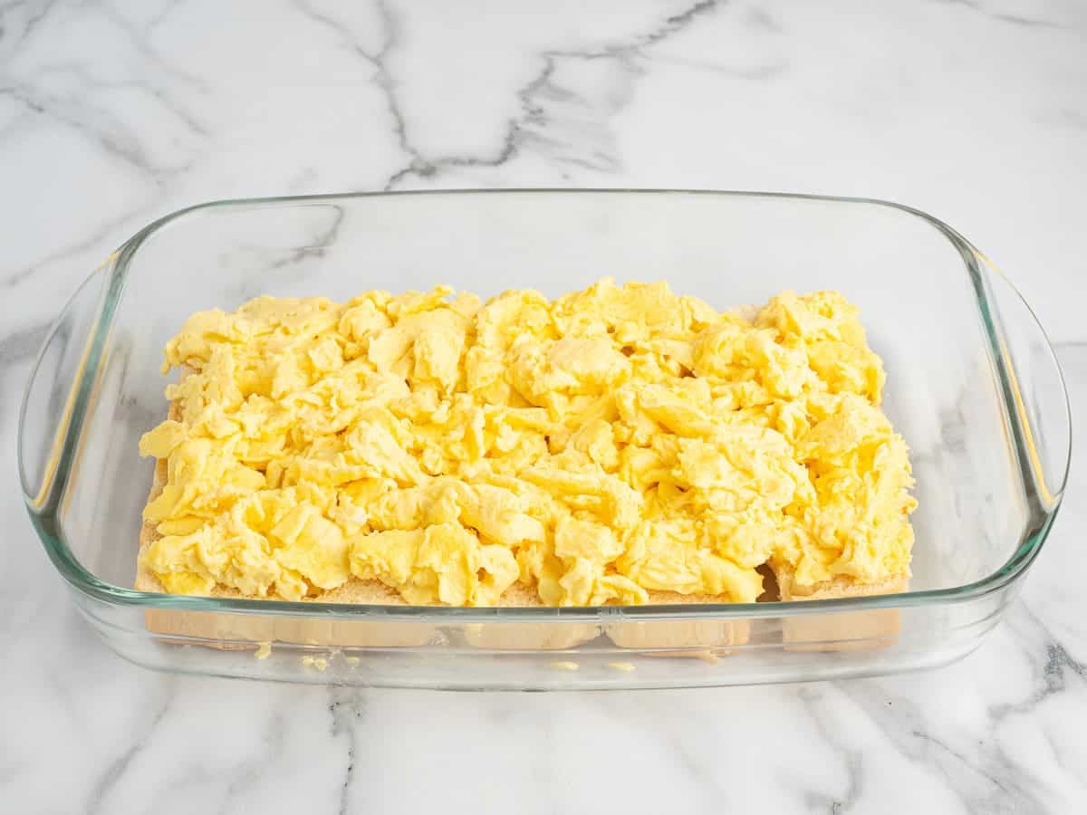 A glass baking dish filled with a layer of scrambled eggs on top of a base of bread pieces, reminiscent of the flavors in Sausage Egg and Cheese Breakfast Sliders, set on a marble countertop.