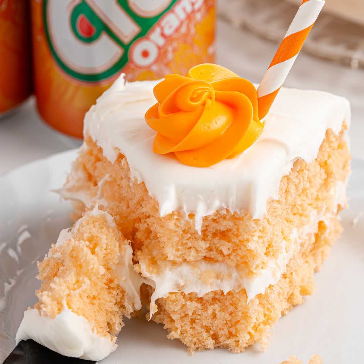 Slice of orange soda cake with a bite removed, topped with a swirl of orange frosting and striped straw, on a white plate.