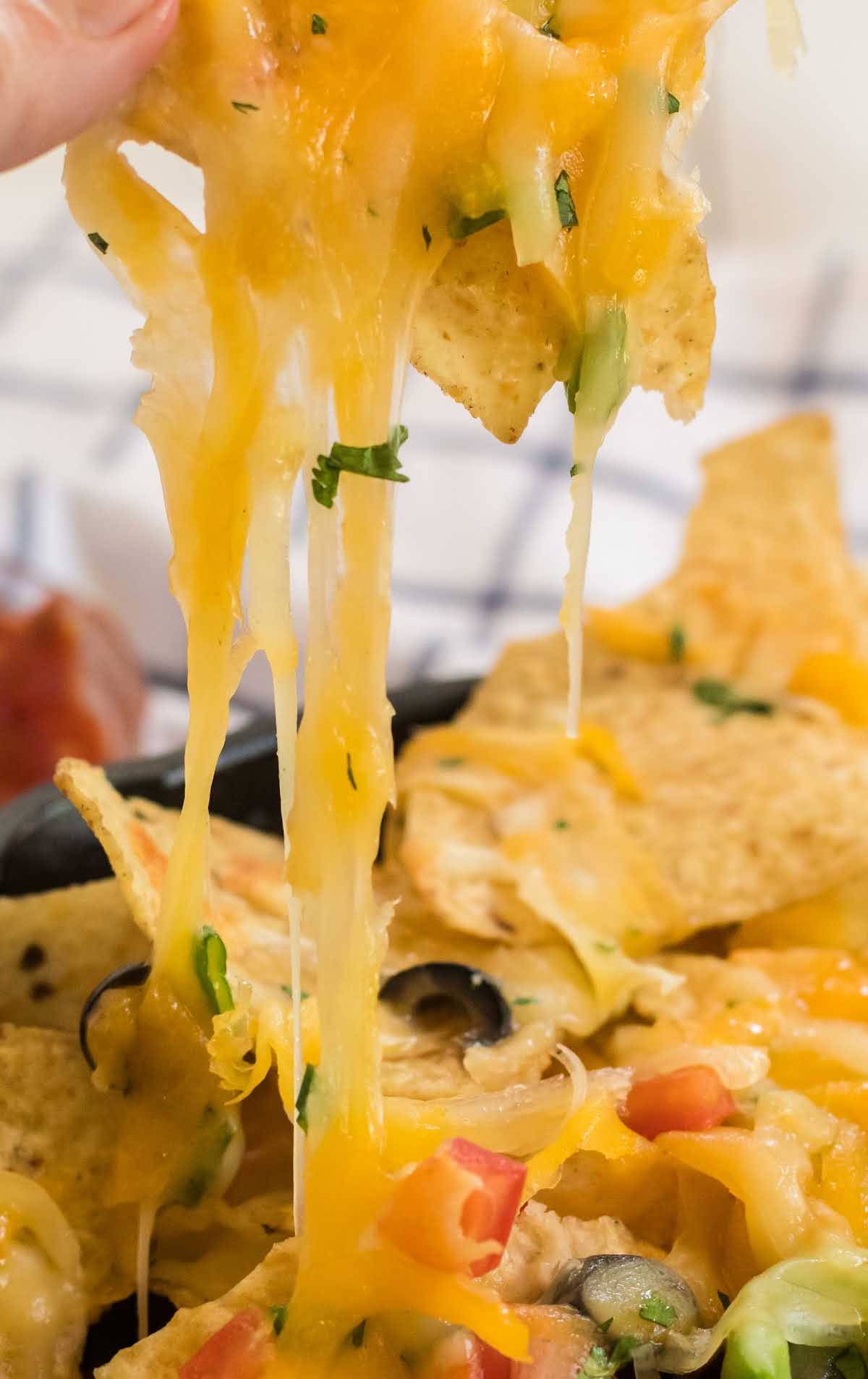 close up shot of nachos being pulled from a skillet of nachos topped with olives, jalapeños, and tomatoes