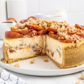 A whole Peach Cobbler Cheesecake is on a serving platter with a few pieces sliced out of it.