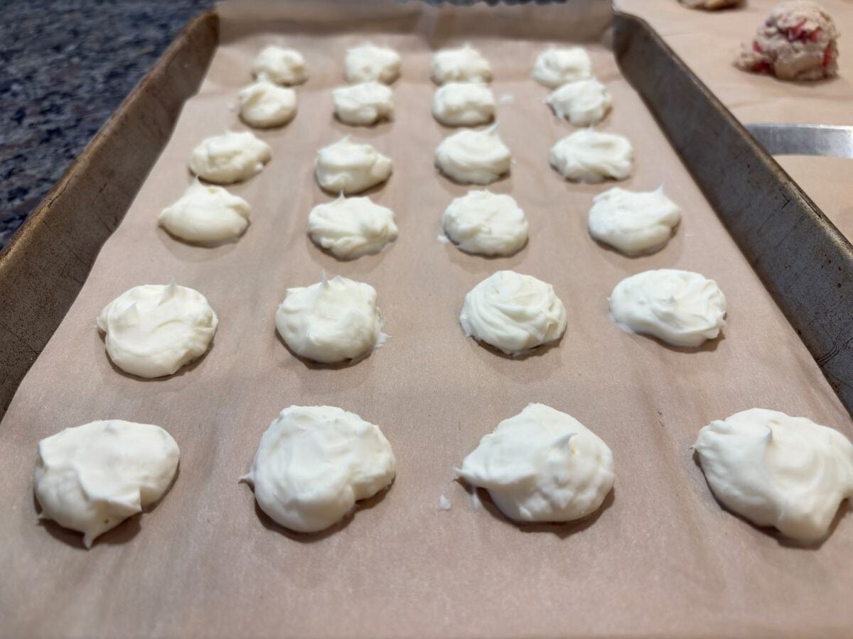 A baking tray lined with parchment paper holds rows of small, round dollops of white Strawberry Cheesecake Cookies dough, ready to be baked. The background shows a countertop and another tray with different dough.