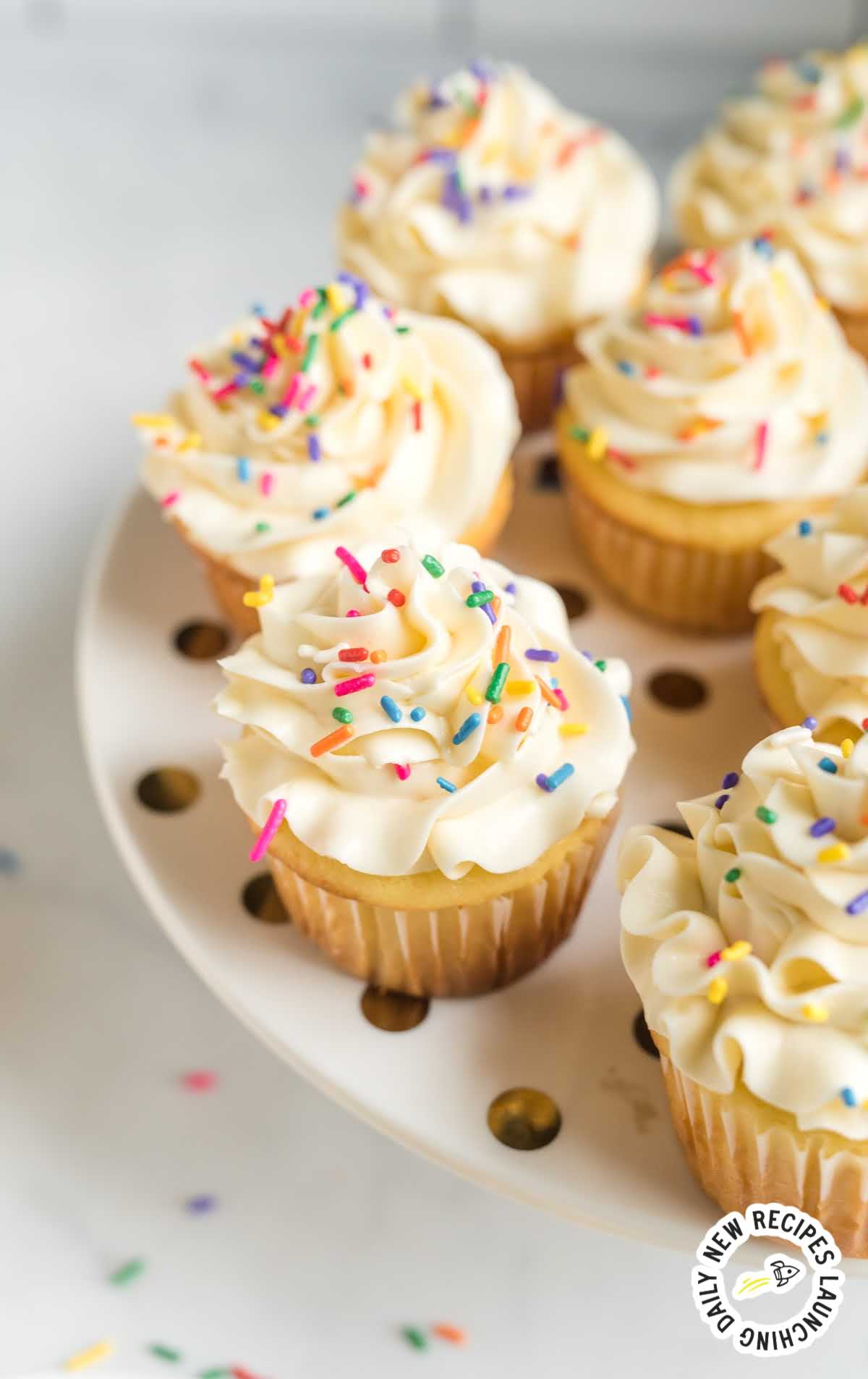 close up shot of a plate of frosted vanilla cupcakes topped with sprinkles