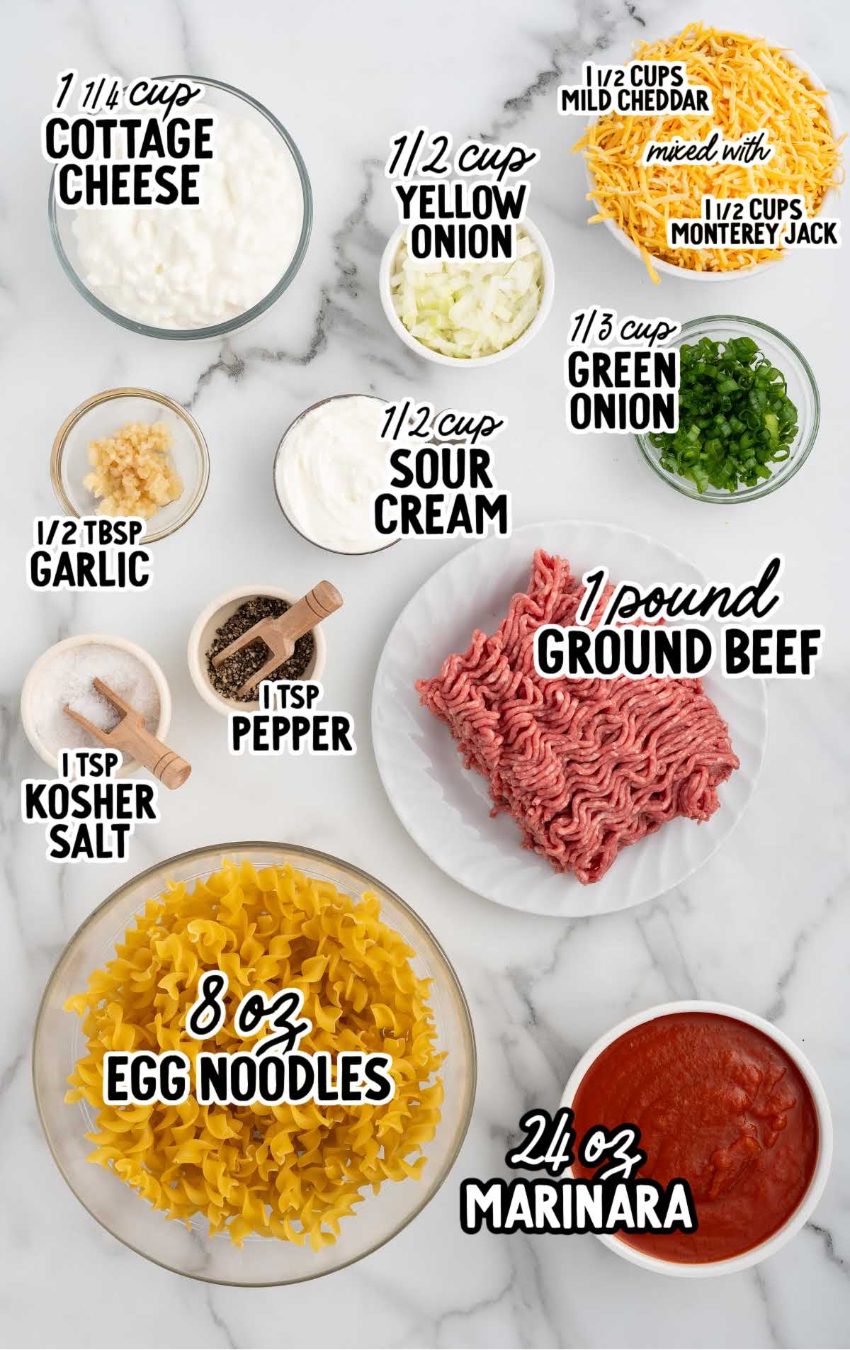 Labeled ingredients for sour cream noodle bake arranged on a marble counter, including egg noodles, ground beef, cheese, cottage cheese, sour cream, onions, marinara, and seasonings.