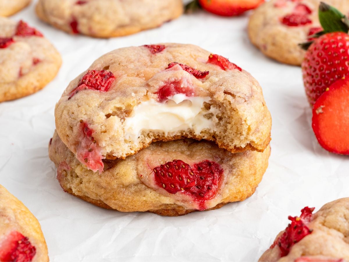 A Strawberry Cheesecake Cookie with a creamy filling is shown with a bite taken out, revealing the inside. Whole and halved strawberries, as well as more Strawberry Cheesecake Cookies, are scattered on white parchment paper.