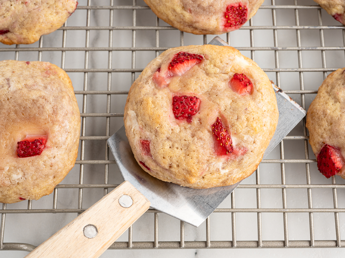 A close-up of freshly baked Strawberry Cheesecake Cookies on a cooling rack. One cookie is being lifted with a metal spatula, revealing chunks of red strawberries and creamy cheesecake swirled into the golden, soft cookie.
