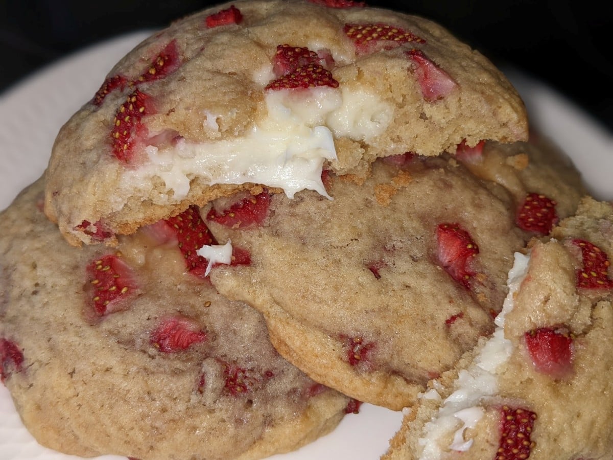 A close-up of Strawberry Cheesecake Cookies filled with white chocolate and chunks of fresh strawberries, with one cookie broken open to reveal the creamy filling inside. The cookies are stacked on a white plate.