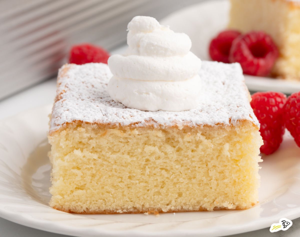 Slice of hot milk cake topped with powdered sugar and a dollop of whipped cream, served on a white plate with fresh raspberries.