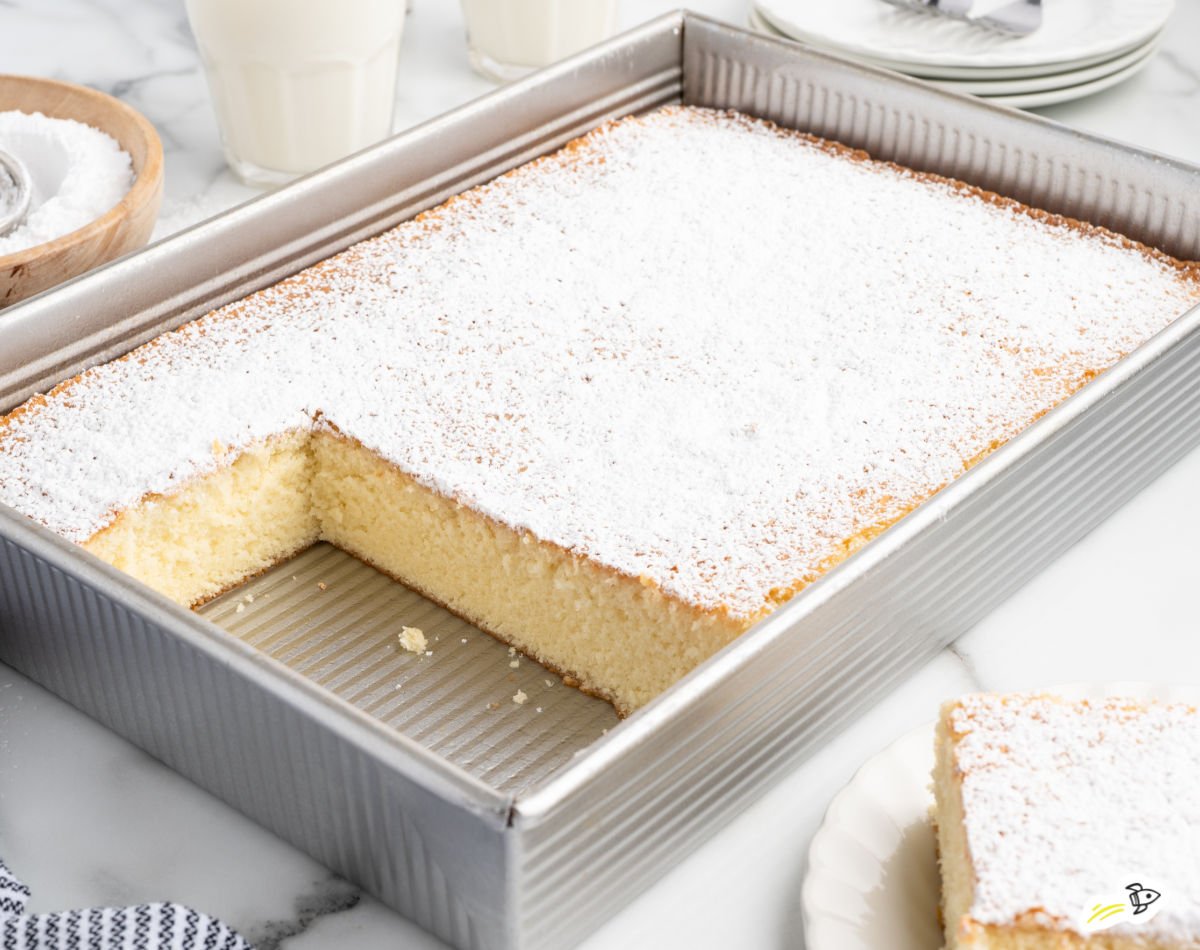 Hot milk cake baked in a metal 9&times;13 pan, dusted generously with powdered sugar, with one square cut out to show the soft vanilla crumb.