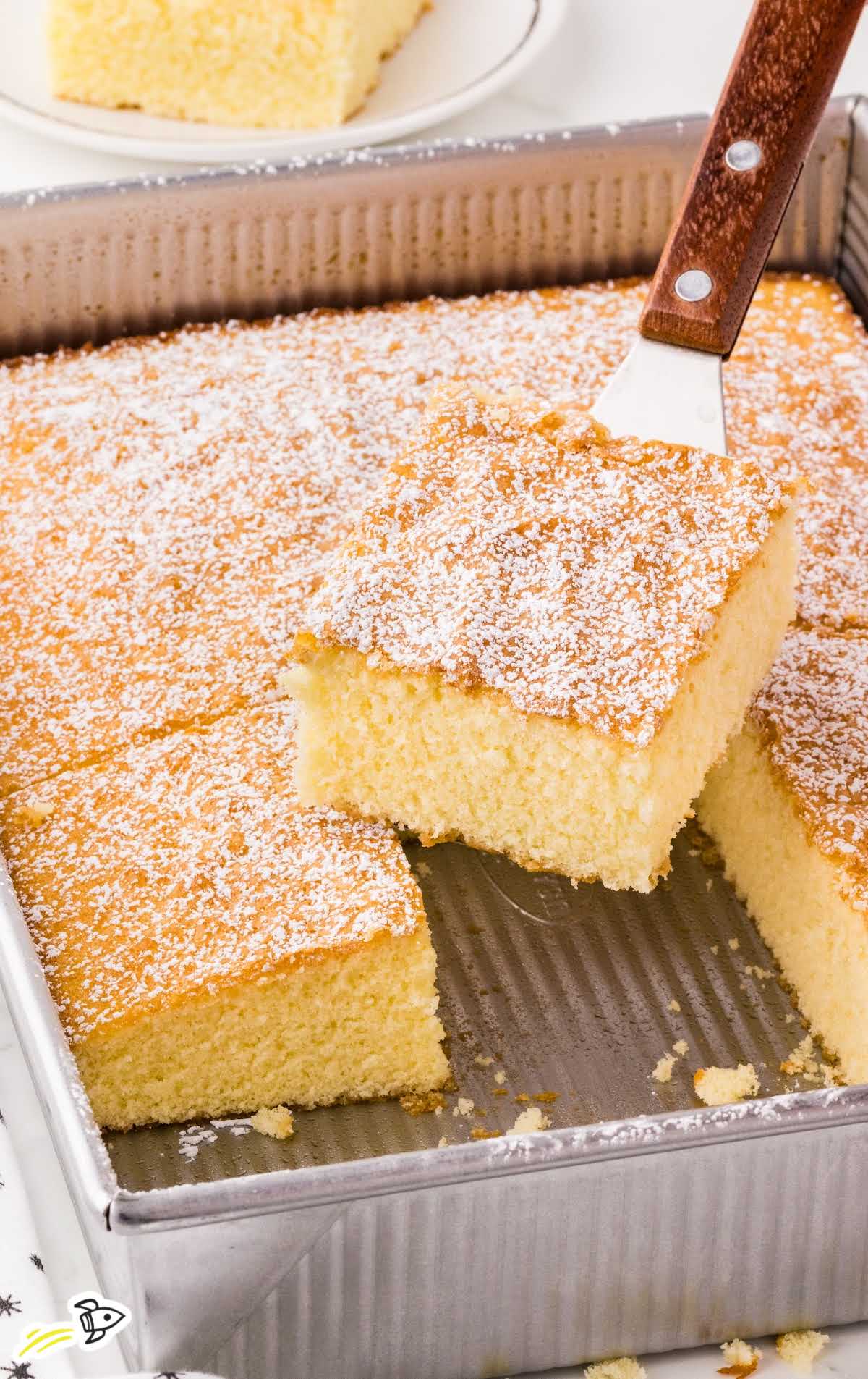 Hot milk cake being lifted from a metal baking pan, showing its soft texture and golden top dusted with powdered sugar.