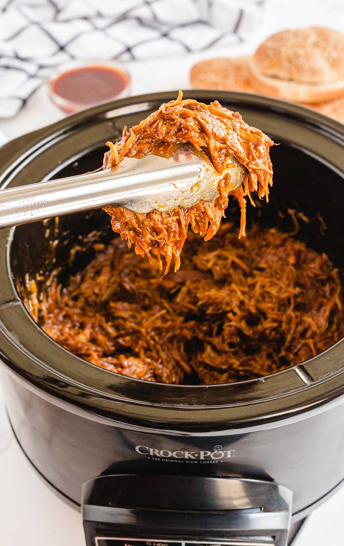 close up shot of pulled bbq chicken in a slow cooker with a piece picked up with tongs