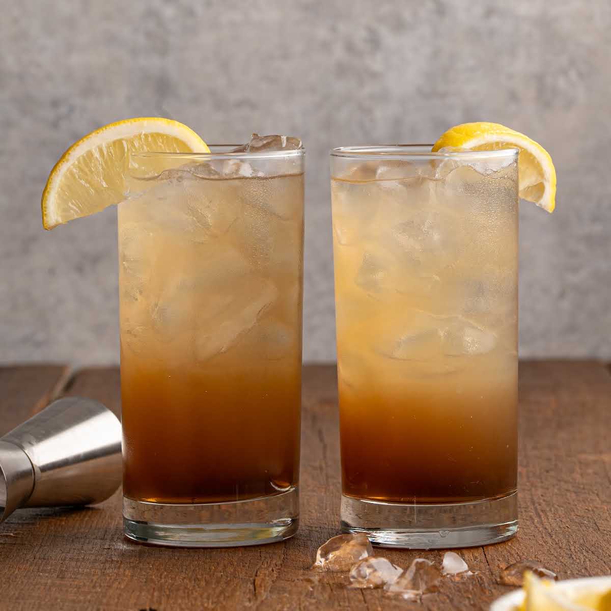 Two glasses of Texas Tea garnished with lemon slices, placed on a wooden surface with a metal jigger nearby.