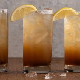 Three tall glasses of Texas Tea with a gradient color effect and lemon wedge garnish on a wooden surface.