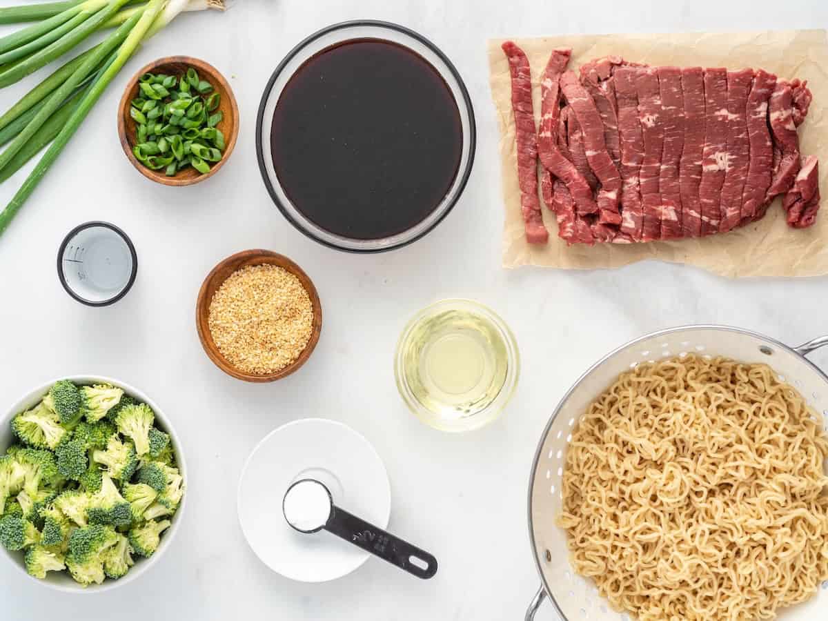Ingredients for a Beef Ramen Stir Fry: sliced beef, uncooked ramen, broccoli, sliced green onions, soy sauce, oil, sesame seeds, black pepper, and salt—all arranged on a white countertop.
