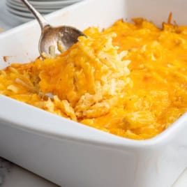 A serving spoon lifts a cheesy Cracker Barrel Hashbrown Casserole from a white baking dish, revealing melted cheddar cheese and shredded potatoes.