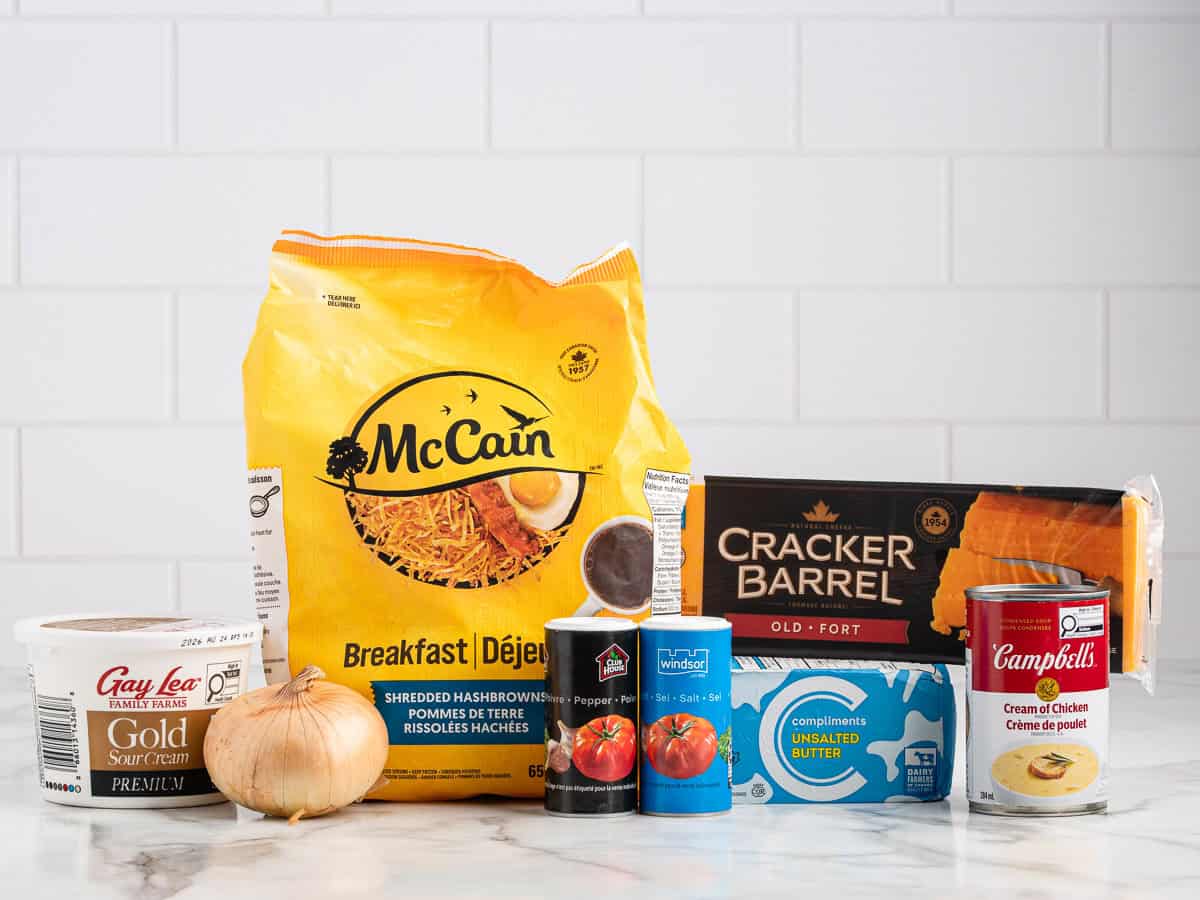 A group of ingredients on a counter — sour cream, onion, bag of hashbrowns, canned diced tomatoes, butter, block of cheddar cheese, and a can of cream of chicken soup — sit ready to be transformed into a comforting Cracker Barrel Hashbrown Casserole against a white tiled background.