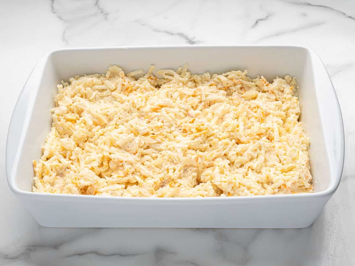 A white baking dish filled with a layer of shredded potatoes and cheese mixture reminiscent of Cracker Barrel Hashbrown Casserole sits on a marble countertop.