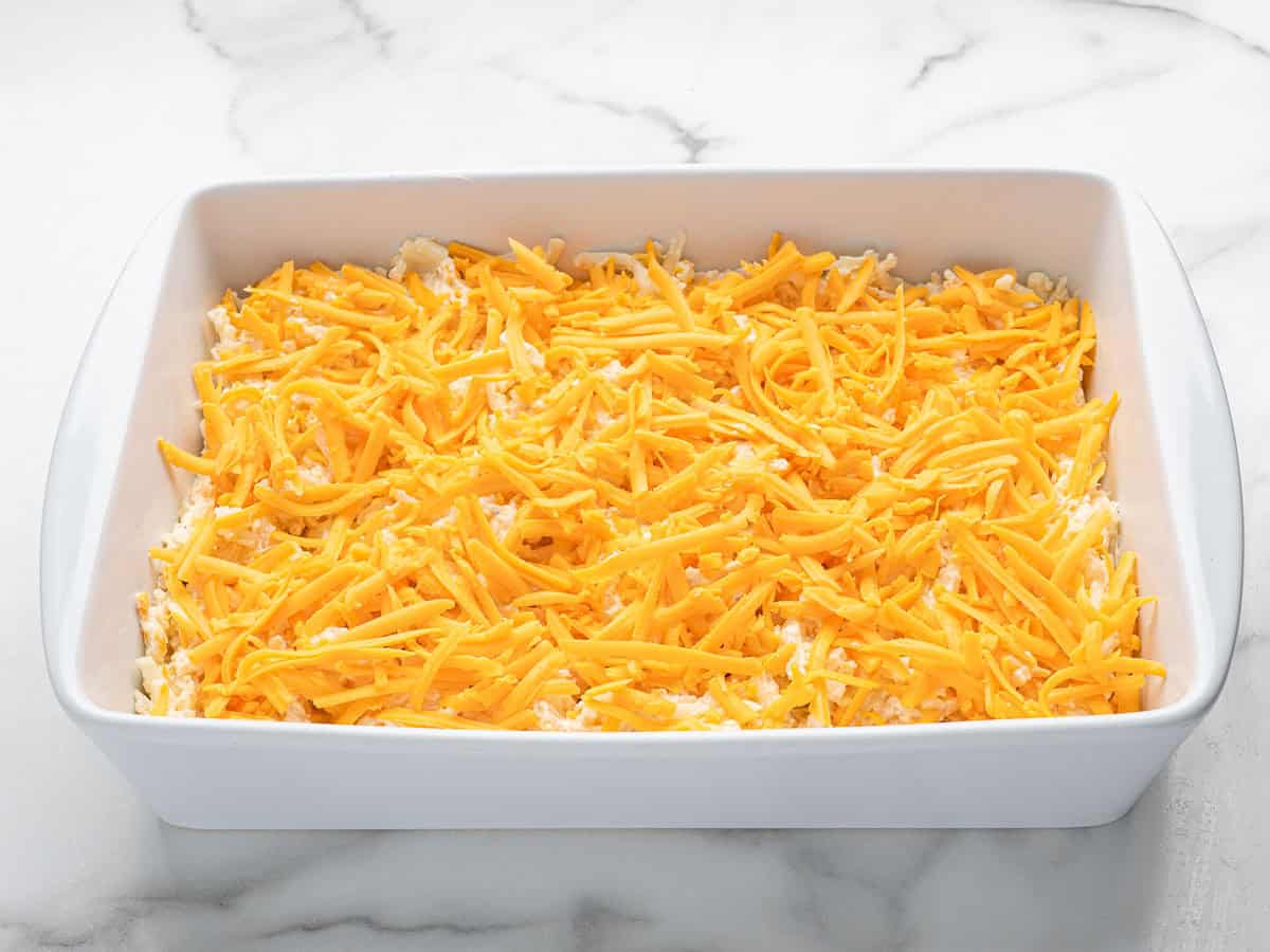 A white baking dish on a marble surface, filled with shredded cheddar cheese evenly spread over a layer of what appears to be a Cracker Barrel Hashbrown Casserole or baked dish base.