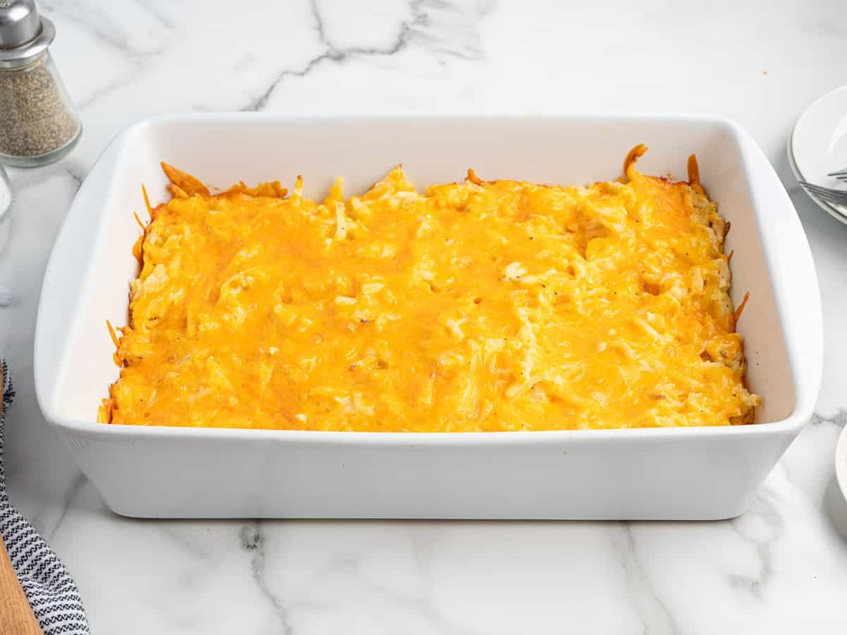 A rectangular white baking dish filled with a golden-brown baked cheesy Cracker Barrel Hashbrown Casserole sits on a marble countertop.