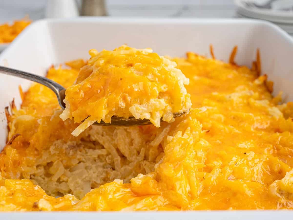 A metal spoon scoops cheesy Cracker Barrel Hashbrown Casserole from a white baking dish, showing melted cheddar cheese on top and creamy, shredded potatoes inside.