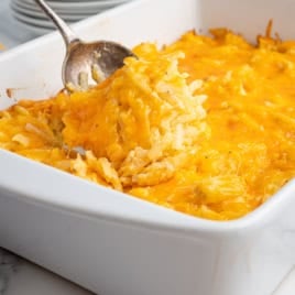 A spoon scoops a cheesy Cracker Barrel Hashbrown Casserole from a white baking dish, showing melted cheddar cheese and shredded potatoes. Plates are stacked in the background.