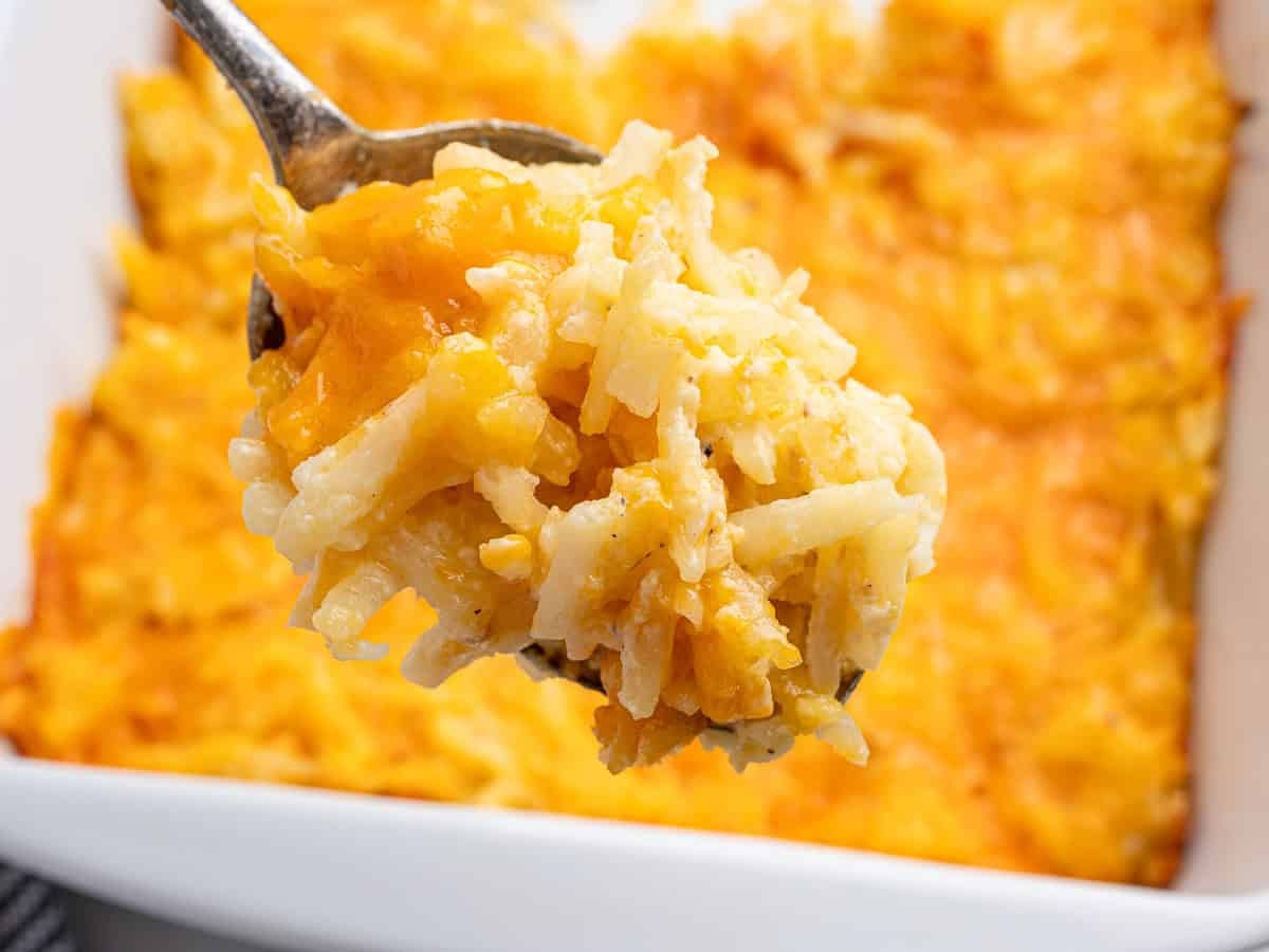 A close-up of a spoonful of cheesy Cracker Barrel Hashbrown Casserole, showing melted cheese and shredded potatoes, held over a white baking dish filled with more of the comforting casserole.