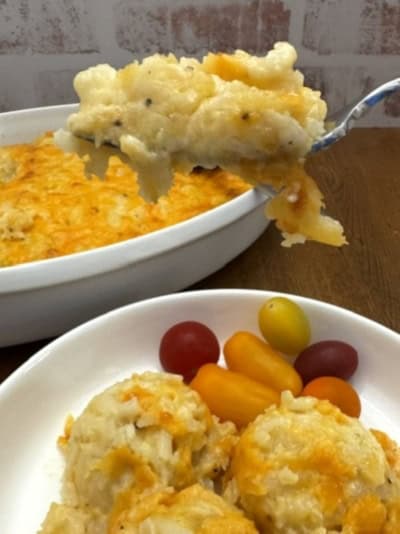 A fork holds a cheesy Cracker Barrel Hashbrown Casserole above a baking dish, with a plate below showing three servings of casserole and colorful grape tomatoes.