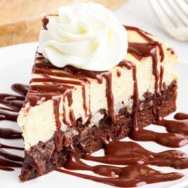 a slice of Brownie Bottom Cheesecake drizzled with chocolate fudge topped with whipped cream