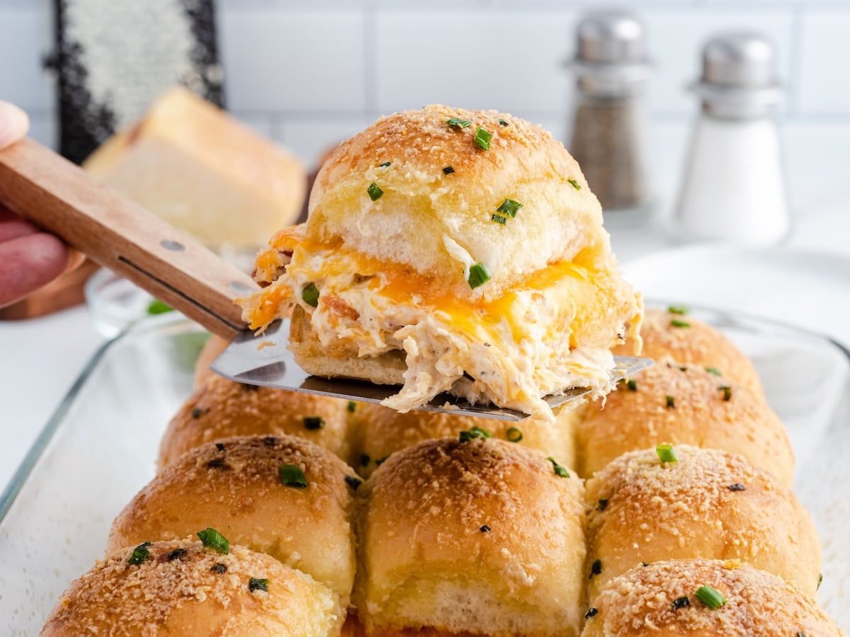 A spatula lifts a cheesy Crack Chicken Slider with golden, toasted buns from a glass baking dish filled with similar sliders. The buns are topped with herbs and cheese. Salt and pepper shakers are in the background.