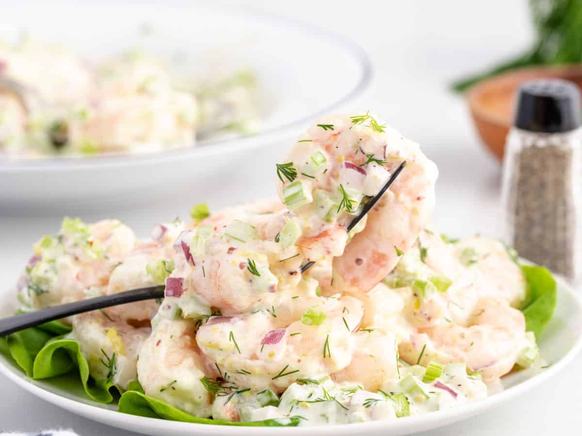 A fork holds up creamy Shrimp Salad with chopped celery, red onion, and dill, served on a bed of lettuce. A pepper shaker and bowl are in the background.