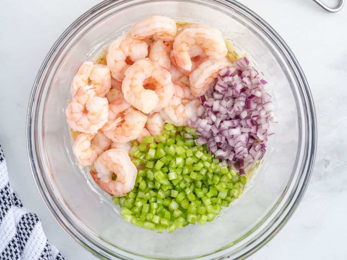 A glass bowl contains the ingredients for a fresh Shrimp Salad—cooked shrimp, diced red onion, and diced celery—each separated into sections. The bowl sits on a white surface next to a black-and-white checkered cloth.