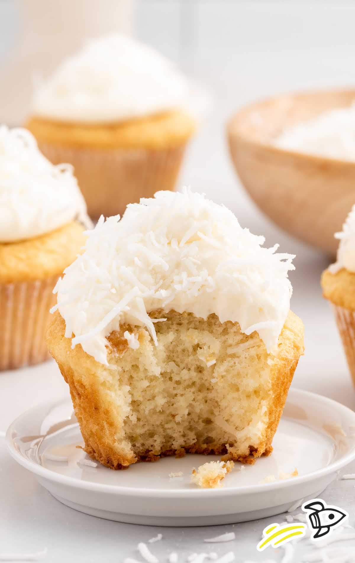 a Coconut Cupcake topped with frosting a coconut flakes with a bite take out on a plate