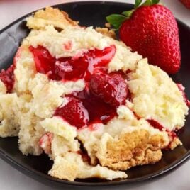 close up shot of a piece of Strawberry Cheesecake Dump Cake on a plate with a strawberry