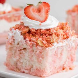 close up shot of a slice of Strawberry Crunch Poke Cake topped with whipped cream and a strawberry on a plate