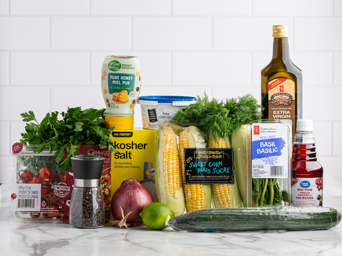 A variety of fresh ingredients for Corn Salad are arranged on a counter&mdash;parsley, grape tomatoes, an onion, cucumber, corn, dill, olive oil, honey, salt, pepper, red wine vinegar, basil, and lime&mdash;set against a white tile background.