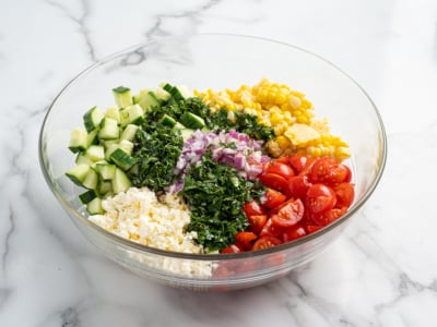 A glass bowl filled with chopped cucumber, cherry tomatoes, corn salad, red onion, fresh herbs, and crumbled feta cheese, all beautifully arranged in separate sections on a white marble surface.
